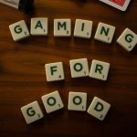 Scrabble tiles on a wooden surface which spell out the words Gaming For Good. There are other board game pieces scattered around them.