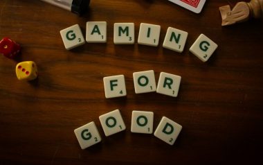 Scrabble tiles on a wooden surface which spell out the words Gaming For Good. There are other board game pieces scattered around them.