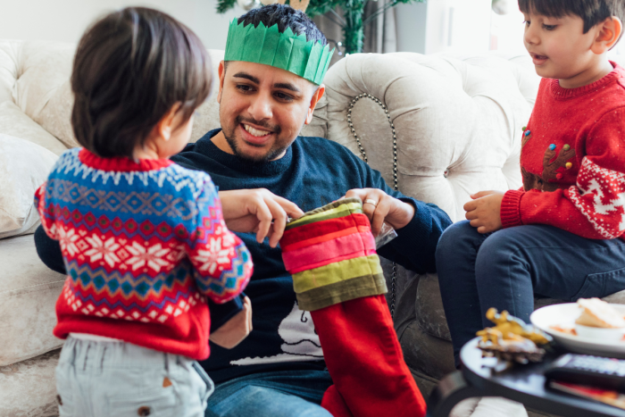 A young person wearing a green paper crown opening a Christmas stocking with two young children who are wearing Christmas jumpers.