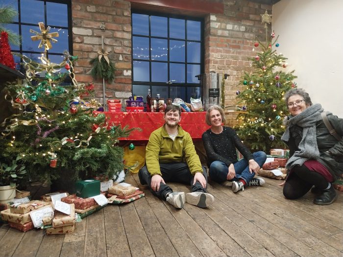 Three people sat next to a Christmas tree with a pile of presents underneath.