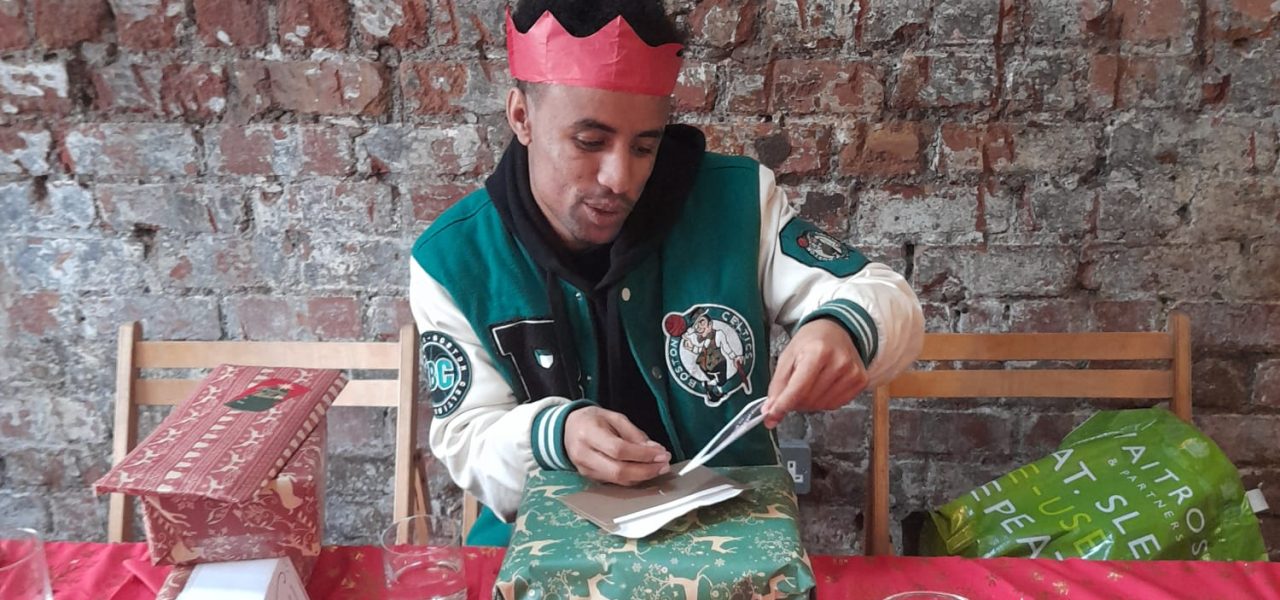 A young person wearing a red paper crown opening a Christmas present at a table with a red table cloth.