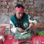 A young person wearing a red paper crown opening a Christmas present at a table with a red table cloth.