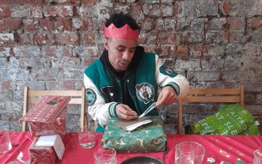 A young person wearing a red paper crown opening a Christmas present at a table with a red table cloth.