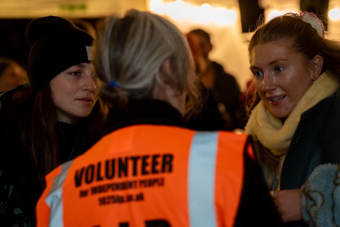 Three people talking one person has their back to us wearing an orange high-vis with he word volunteer in black writing.